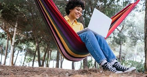 一位在线学生坐在树下的红色条纹吊床上学习他们的在线课程. 